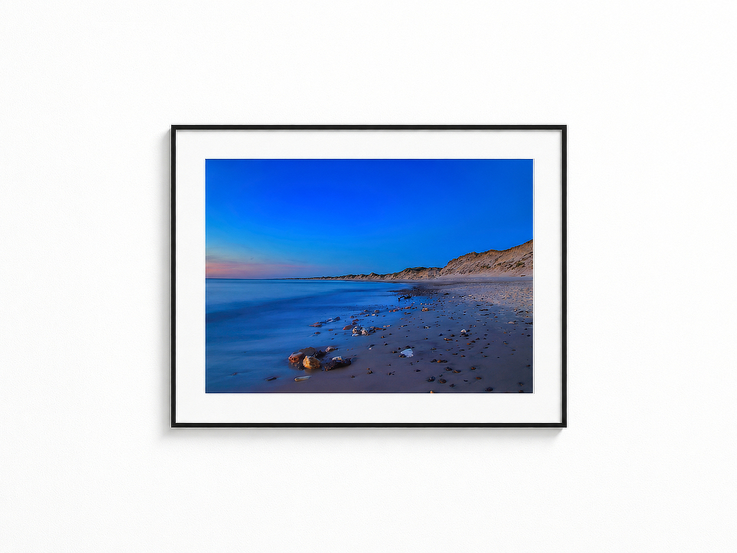 Visualisierung der Fotografie „Endlosblau“ in schwarzem Rahmen mit weißem Passepartout an einer hellen Wand. Zu sehen ist ein weiter Strand mit ruhigem Meer, vereinzelten Steinen und einer Dünenküste unter tiefblauem Abendhimmel.