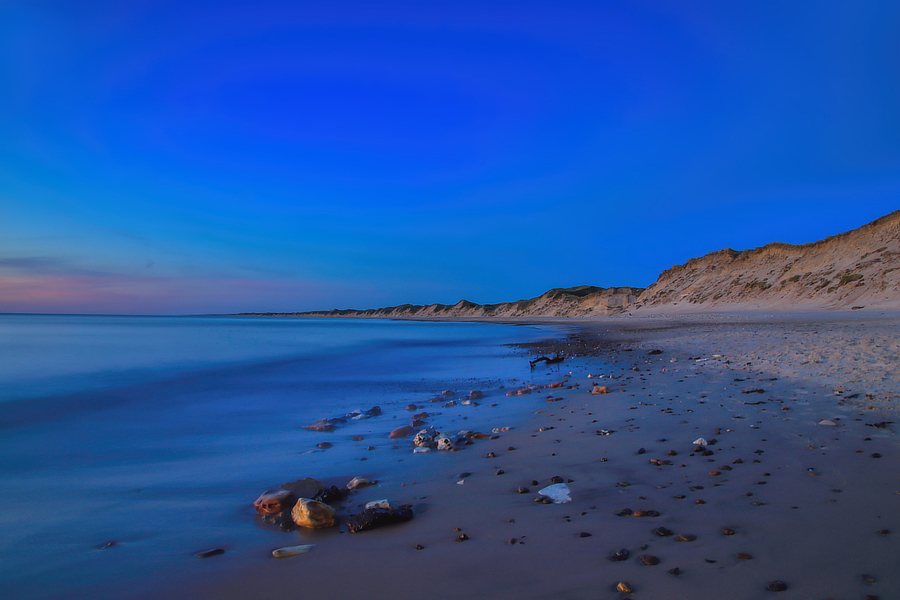 Blick entlang eines weiten Strandes mit ruhigem Meer, vereinzelten Steinen und Dünenküste unter tiefblauem Abendhimmel.