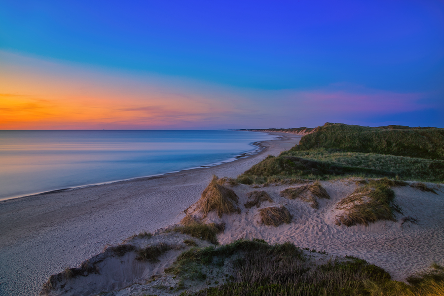 Blick über Dünen und einen weiten Strand auf ein ruhig wirkendes Meer unter blau-orangefarbenem Abendhimmel.