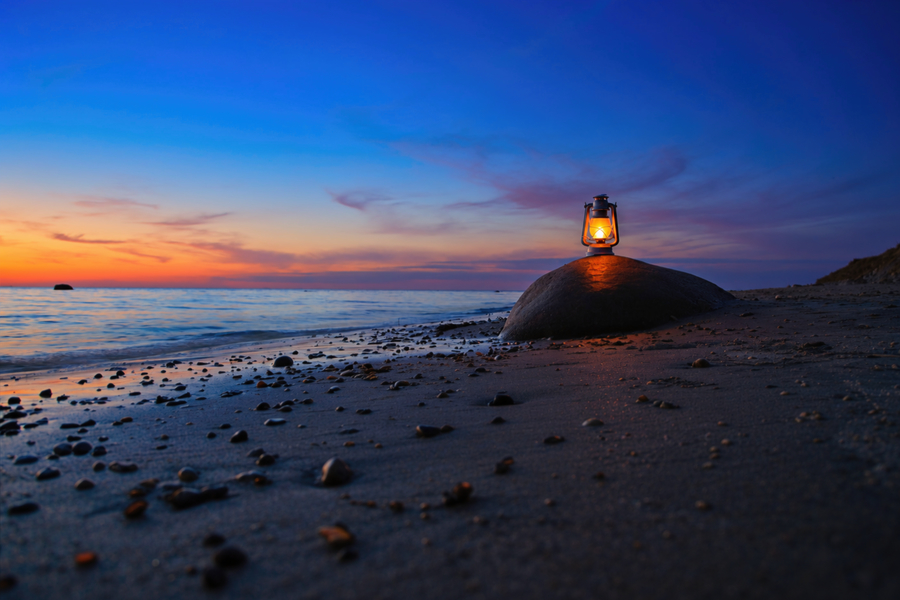 Leuchtende Laterne auf einem großen Stein am Strand vor ruhigem Meer und farbigem Abendhimmel.