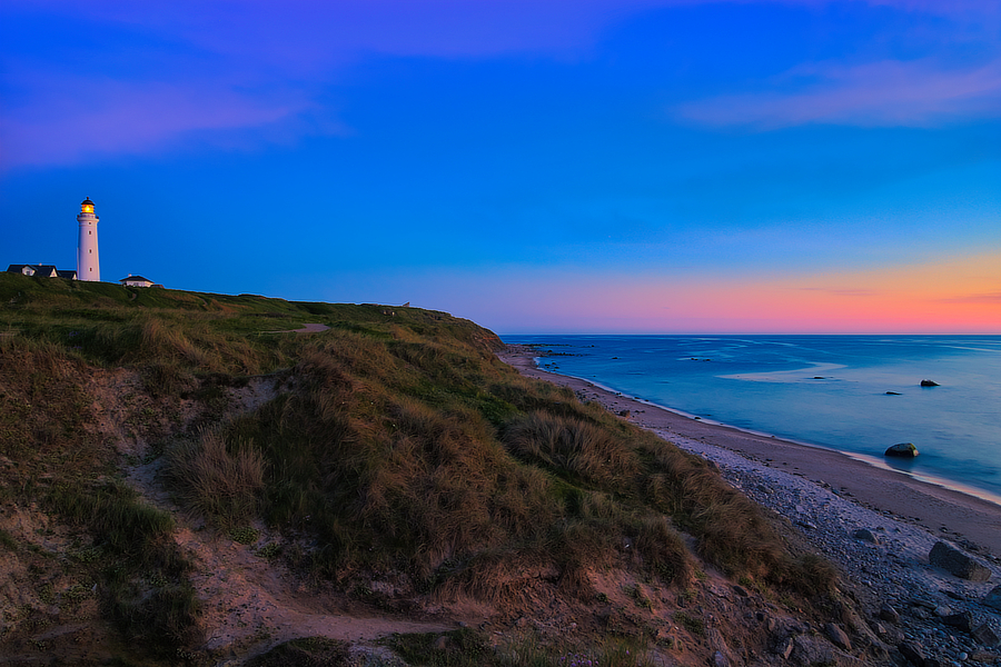 Leuchtturm auf einer grasbewachsenen Steilküste über dem ruhigen Meer bei blau-rosa Abendhimmel.