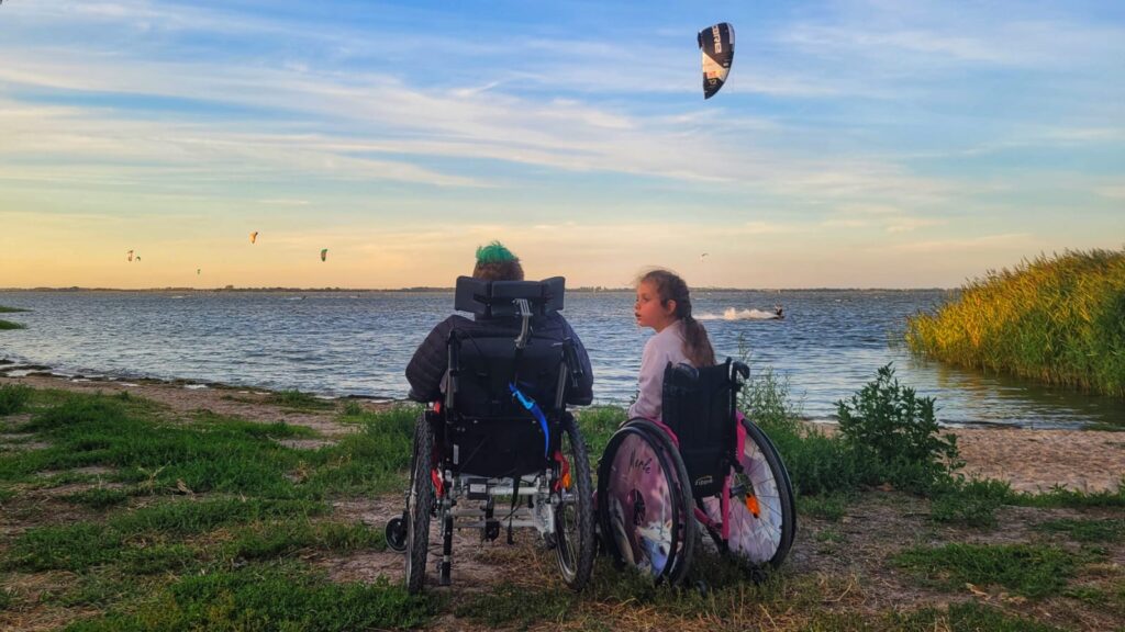 Zwei junge Menschen im Rollstuhl sitzen nebeneinander am Ufer und blicken über das Wasser. Über ihnen stehen mehrere Kites im weiten Abendhimmel.