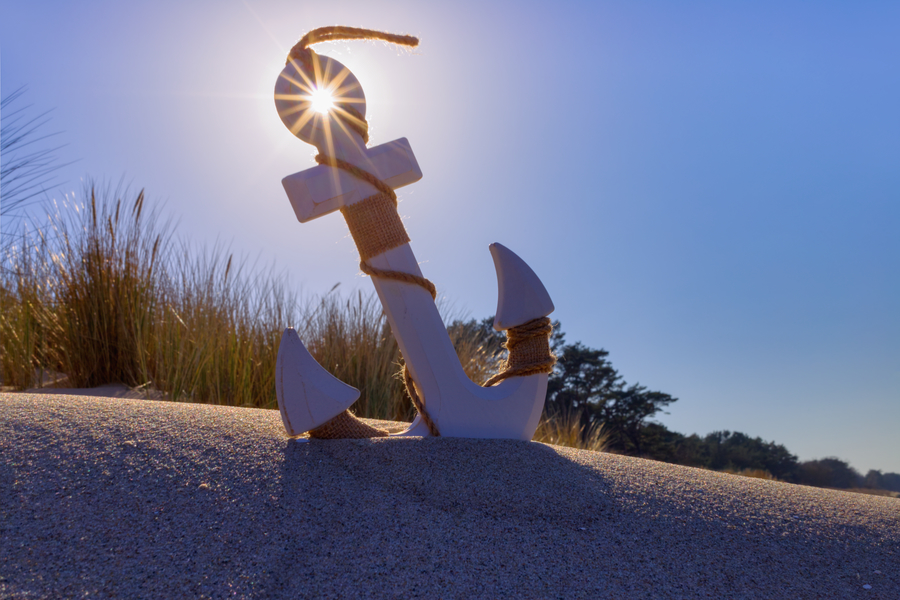 Weißer dekorativer Anker steht im Sand einer Düne vor hohem Dünengras und blauem Himmel. Die tief stehende Sonne scheint durch die obere Öffnung des Ankers und bildet einen deutlich sichtbaren Blendenstern.