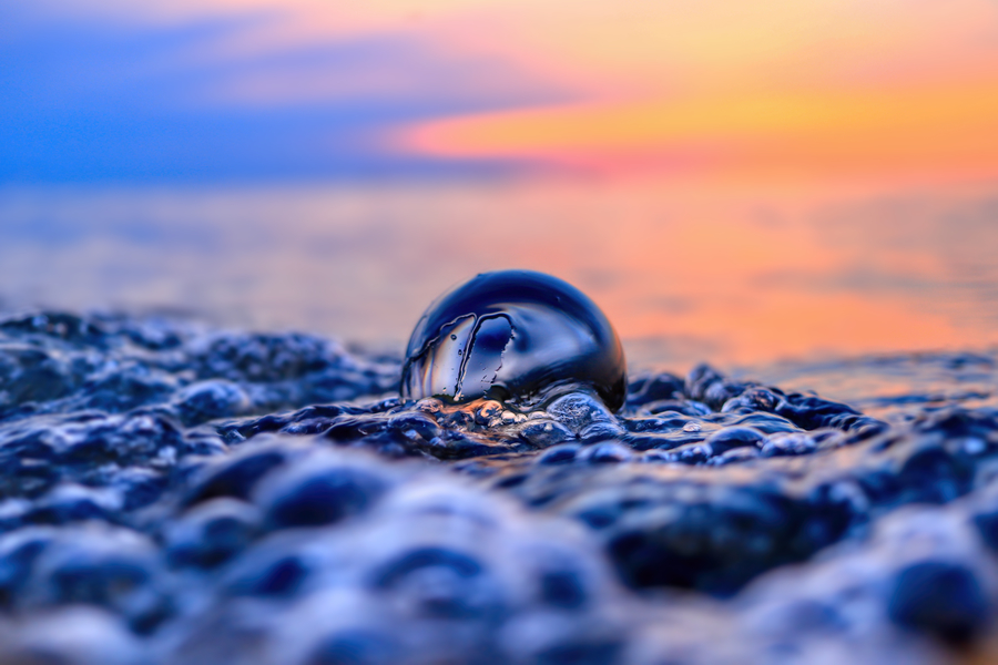 Klare Glaskugel wie ein großer Wassertropfen zwischen nassen Steinen am Ufer, vor weich leuchtendem orange-blauem Abendhimmel.