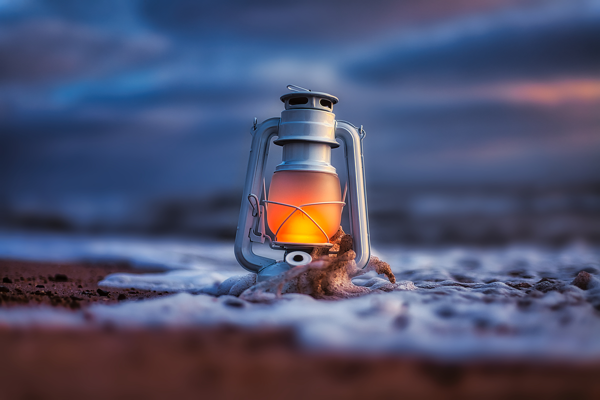 Warm leuchtende Laterne am Strand, von einer heranlaufenden Welle und weißem Schaum umspült, vor dunkelblauem Abendhimmel.