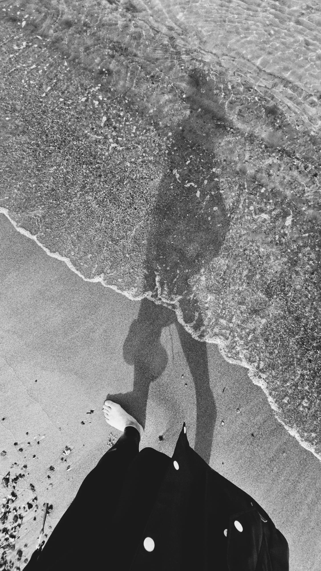 L I C H T E R M E E R – Meerwärts chwarz-Weiß-Aufnahme von nackten Füßen am Strand, während eine schäumende Welle über den Sand und die Füße läuft.