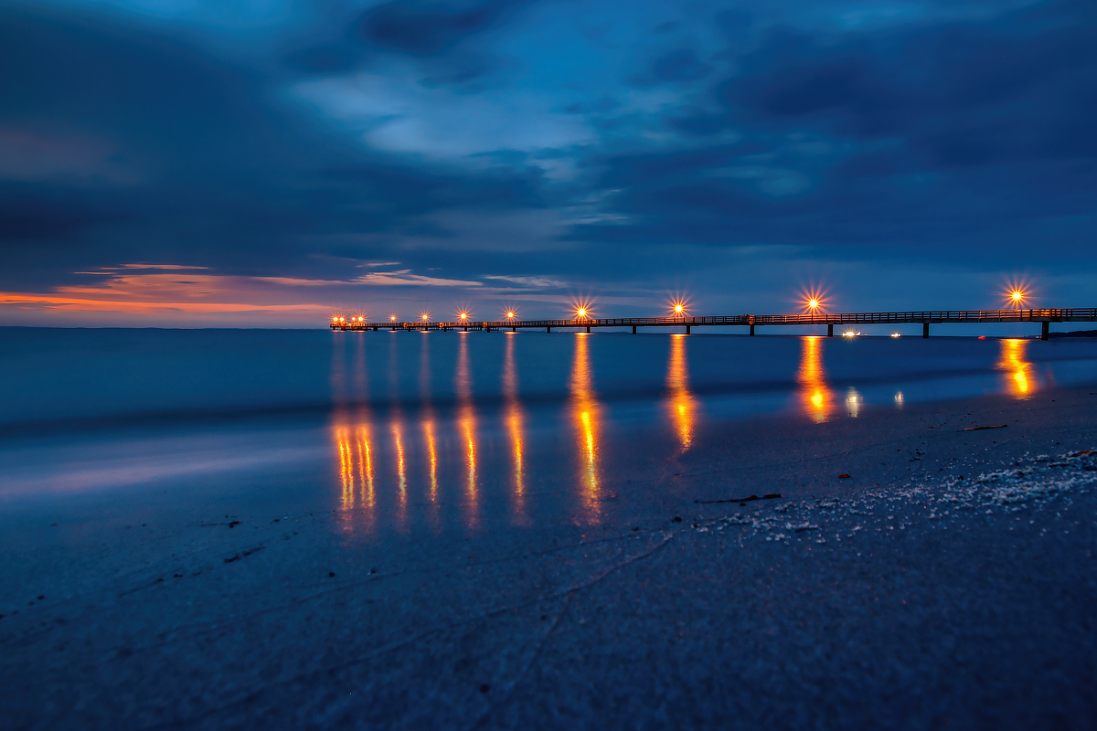 Beleuchtete Seebrücke in der blauen Stunde mit sternförmigen Lichtern und warmen Spiegelungen auf dem ruhigen Meer.