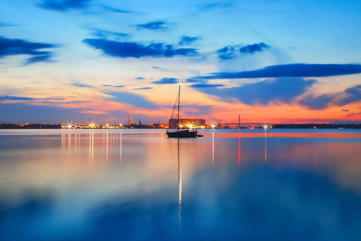 Segelboot auf spiegelglattem Wasser in der blauen Stunde mit leuchtenden Hafenlichtern, Blendensternen und langen Spiegelungen.