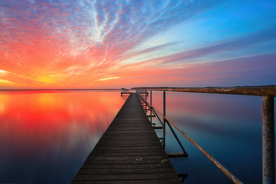 Langer Holzsteg über spiegelglattem Wasser vor leuchtend orange-blauem Abendhimmel mit intensiver Spiegelung der Farben im Meer.