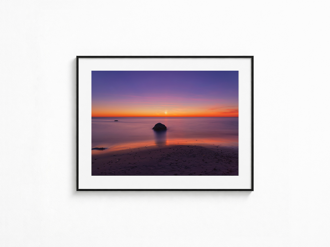 Visualisierung der Fotografie „Unendlicht“ in schwarzem Rahmen mit weißem Passepartout an einer hellen Wand. Langzeitbelichtung eines ruhigen Meeres mit einzelnen Felsen vor leuchtend orange-violettem Abendhimmel.