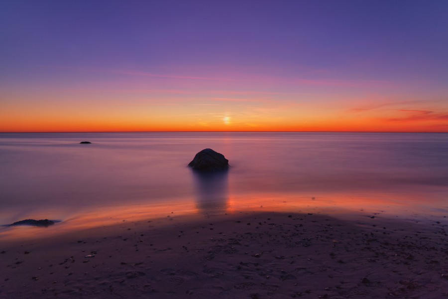Langzeitbelichtung eines ruhigen Meeres mit einzelnen Felsen vor leuchtend orange-violettem Abendhimmel.