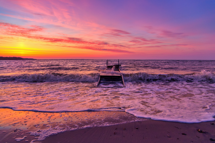 Alter zerstörter Steg in der Brandung vor leuchtend orange-rosa Sonnenuntergang, während Wellen auf den Strand laufen.
