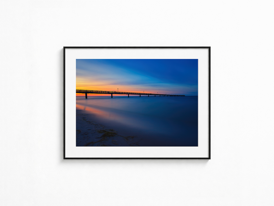 Visualisierung der Fotografie „Wogentänzer“ in schwarzem Rahmen mit weißem Passepartout an einer hellen Wand. Lange Seebrücke über ruhig wirkendem Meer vor orange leuchtendem Sonnenuntergang und tiefblauem Abendhimmel.