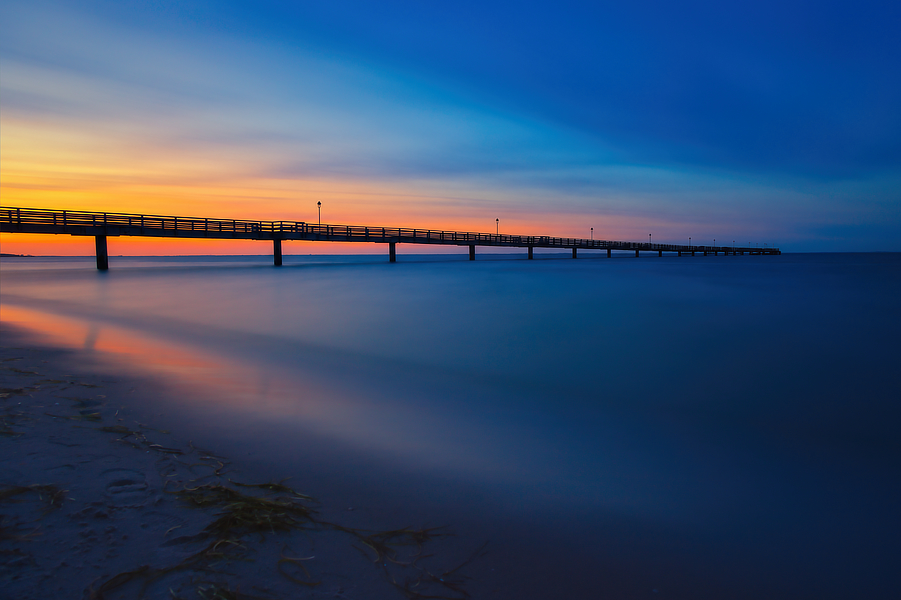 Lange Seebrücke über ruhig wirkendem Meer vor orange leuchtendem Sonnenuntergang und tiefblauem Abendhimmel.
