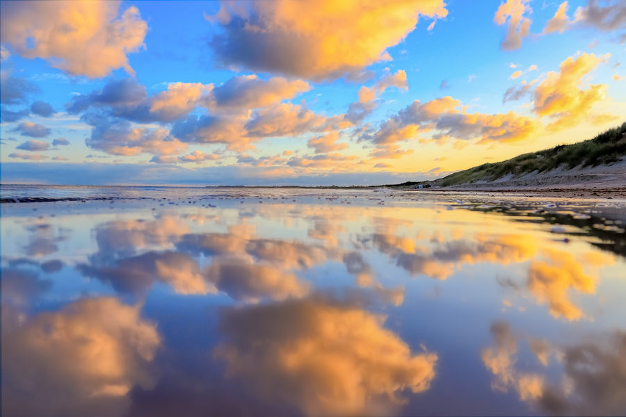 Am Strand spiegeln sich große orange leuchtende Wolken und der blaue Himmel im ruhigen Wasser, während rechts eine grasbewachsene Düne entlang der Küste verläuft.