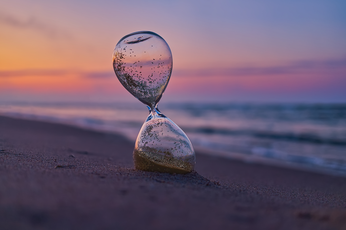Gläserne Sanduhr mit glitzerndem Sand am Strand vor unscharfem Meer und pastellfarbenem Abendhimmel.