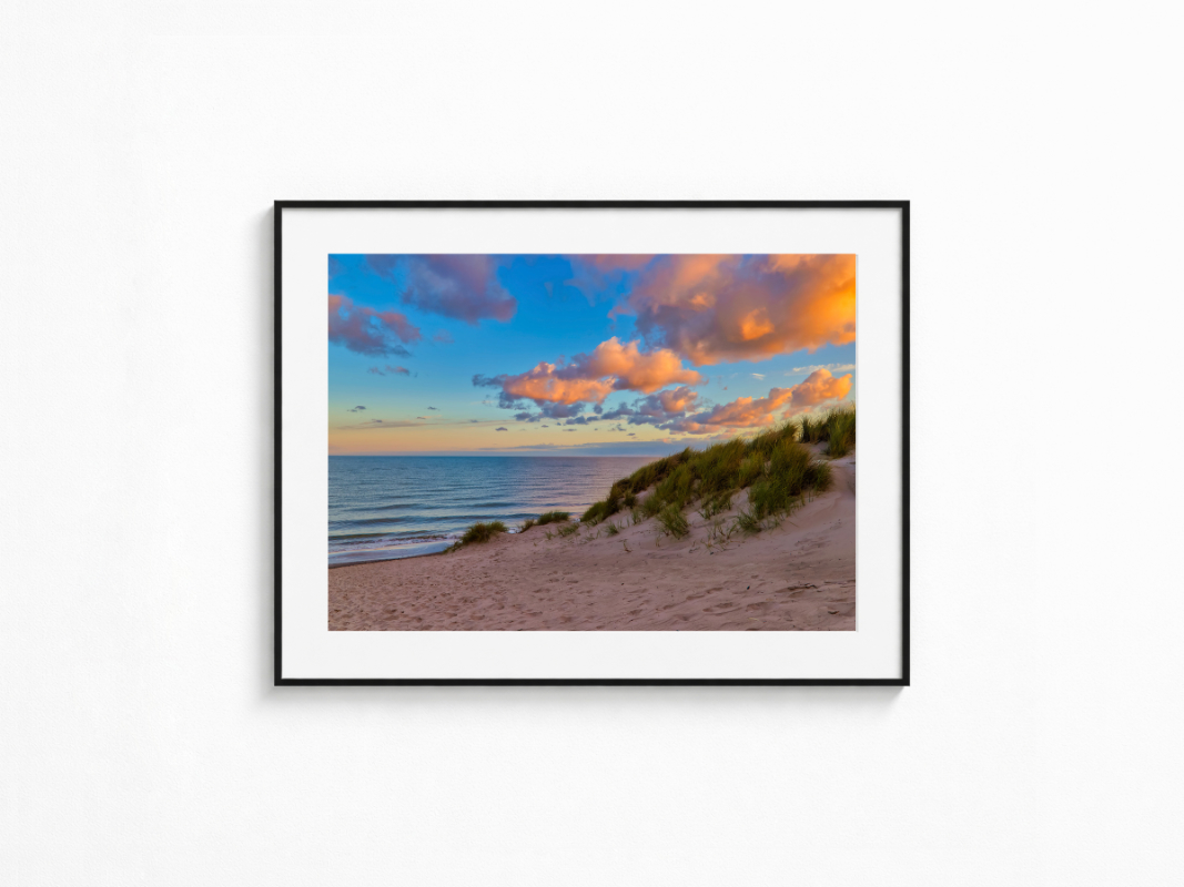 Visualisierung der Fotografie „Zwischenlicht“ in schwarzem Rahmen mit weißem Passepartout an einer hellen Wand. Blick über eine sandige Düne mit hohem Dünengras auf das ruhige Meer. Große Wolken über der Küste werden von warmem orangefarbenem Abendlicht angestrahlt, während der Himmel bereits tiefblau wird.