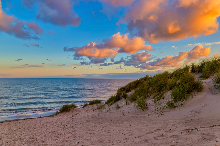 Blick über eine sandige Düne mit hohem Dünengras auf das ruhige Meer. Große Wolken über der Küste werden von warmem orangefarbenem Abendlicht angestrahlt, während der Himmel bereits tiefblau wird.