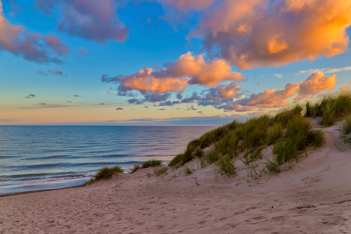 Blick über eine sandige Düne mit hohem Dünengras auf das ruhige Meer. Große Wolken über der Küste werden von warmem orangefarbenem Abendlicht angestrahlt, während der Himmel bereits tiefblau wird.