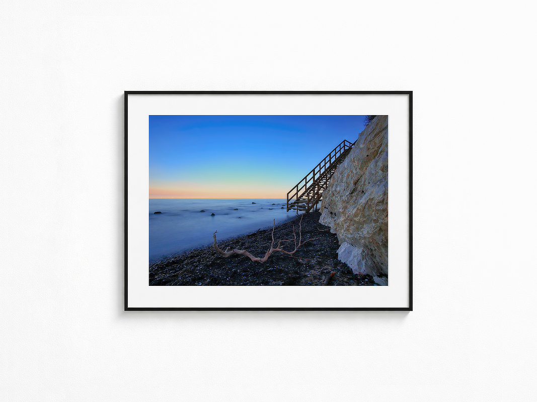 Visualisierung der Fotografie „Blaupause“ in schwarzem Rahmen mit weißem Passepartout an einer hellen Wand. Langzeitbelichtung an einer Steilküste mit Treppe zum Meer in der blauen Stunde.