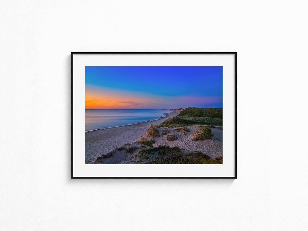 Visualisierung der Fotografie „Ewigkeitsrausch“ in schwarzem Rahmen mit weißem Passepartout an einer hellen Wand. Blick über Dünen und einen weiten Strand auf ein ruhig wirkendes Meer unter blau-orangefarbenem Abendhimmel.