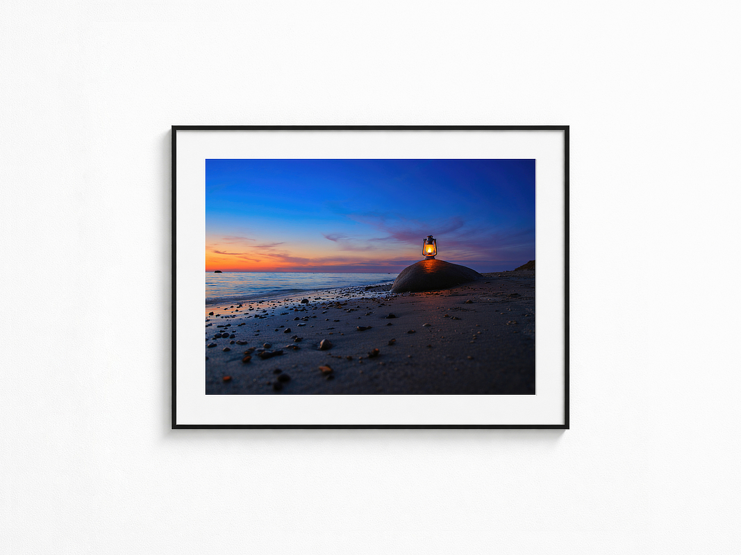 Visualisierung der Fotografie „Glücksfunke“ in schwarzem Rahmen mit weißem Passepartout an einer hellen Wand. Leuchtende Laterne auf einem großen Stein am Strand vor ruhigem Meer und farbigem Abendhimmel.