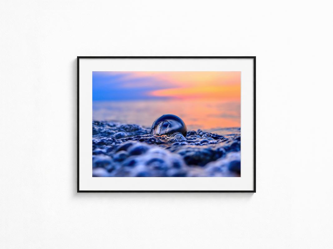 Visualisierung der Fotografie „Lichthauch“ in schwarzem Rahmen mit weißem Passepartout an einer hellen Wand. Gläserne Kugel zwischen Wasser und Steinen im weichen Abendlicht.