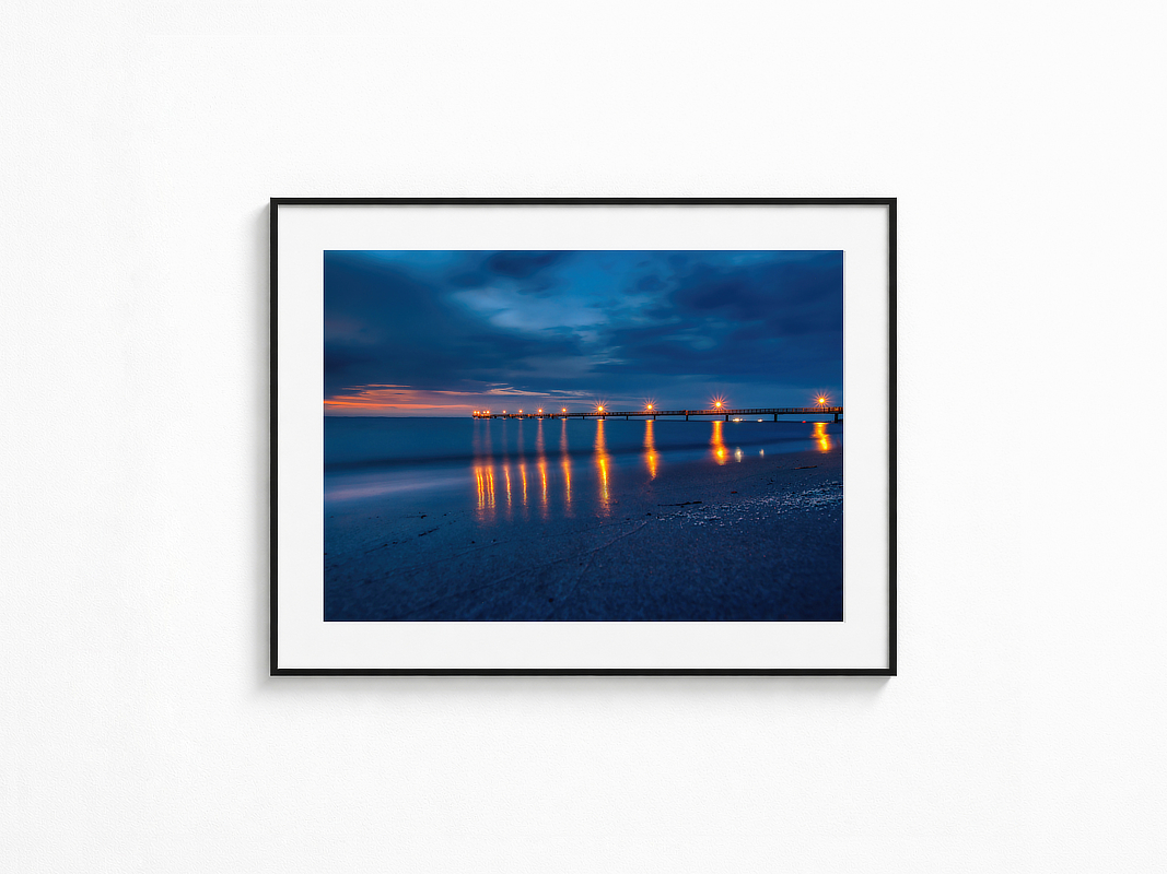 Visualisierung der Fotografie „Nirgendwohin“ in schwarzem Rahmen mit weißem Passepartout an einer hellen Wand. Beleuchtete Seebrücke in der blauen Stunde mit sternförmigen Lichtern und warmen Spiegelungen auf dem ruhigen Meer.