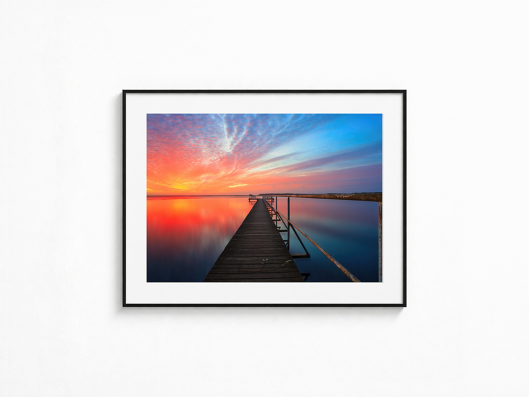 Visualisierung der Fotografie „Traumtänzer“ in schwarzem Rahmen mit weißem Passepartout an einer hellen Wand. Langer Holzsteg über spiegelglattem Wasser vor leuchtend orange-blauem Abendhimmel mit intensiver Spiegelung der Farben im Meer.