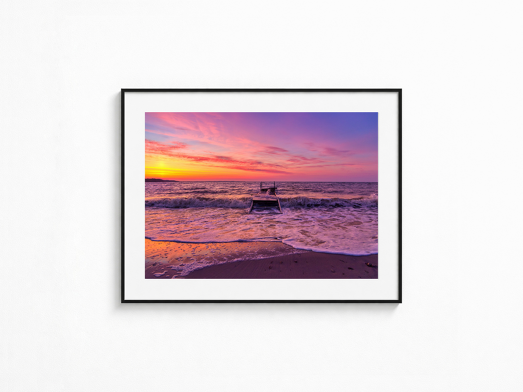 Visualisierung der Fotografie „Wellenreigen“ in schwarzem Rahmen mit weißem Passepartout an einer hellen Wand. Alter Steg in der Brandung vor leuchtend orange-rosa Sonnenuntergang, während Wellen auf den Strand laufen.