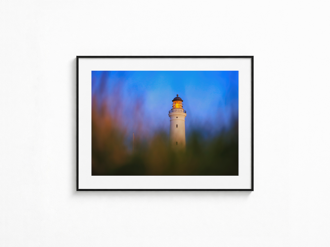 Visualisierung der Fotografie „Wellenstille“ in schwarzem Rahmen mit weißem Passepartout an einer hellen Wand. Leuchtturm mit warm leuchtender Laterne in der blauen Stunde, gesehen durch weich unscharfe Gräser im Vordergrund.
