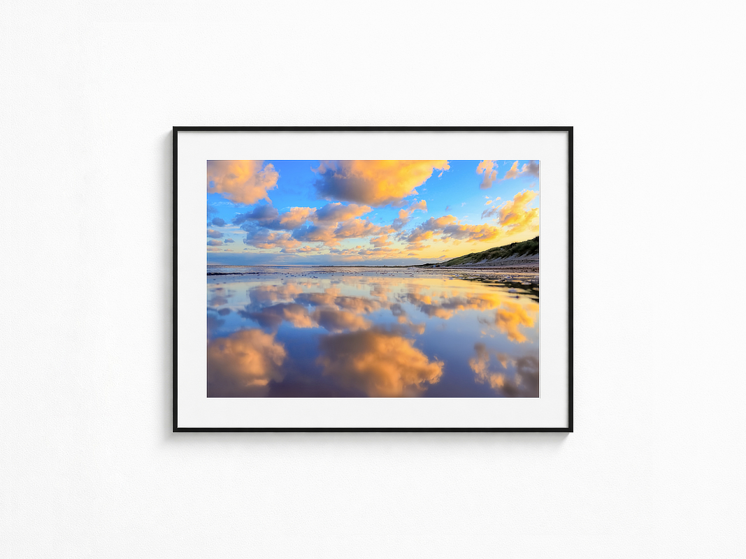 Visualisierung der Fotografie „Wolkenmeer“ in schwarzem Rahmen mit weißem Passepartout an einer hellen Wand. Am Strand spiegeln sich große orange leuchtende Wolken und der blaue Himmel im ruhigen Wasser, während rechts eine grasbewachsene Düne entlang der Küste verläuft.